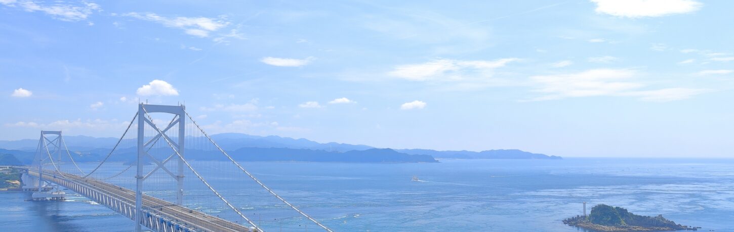 A high-angle view of the magnificent Akashi Kaikyo Bridge stretching across the bright blue sea toward Awaji Island under a clear sky.
