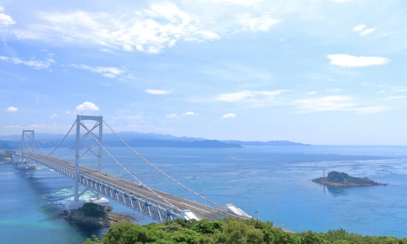 A high-angle view of the magnificent Akashi Kaikyo Bridge stretching across the bright blue sea toward Awaji Island under a clear sky.