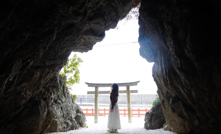 A person standing inside a rocky cave, looking out toward a traditional stone torii gate and the sea on Awaji Island.