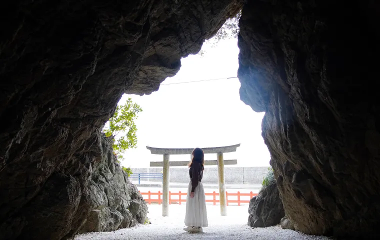 A person standing inside a rocky cave, looking out toward a traditional stone torii gate and the sea on Awaji Island.