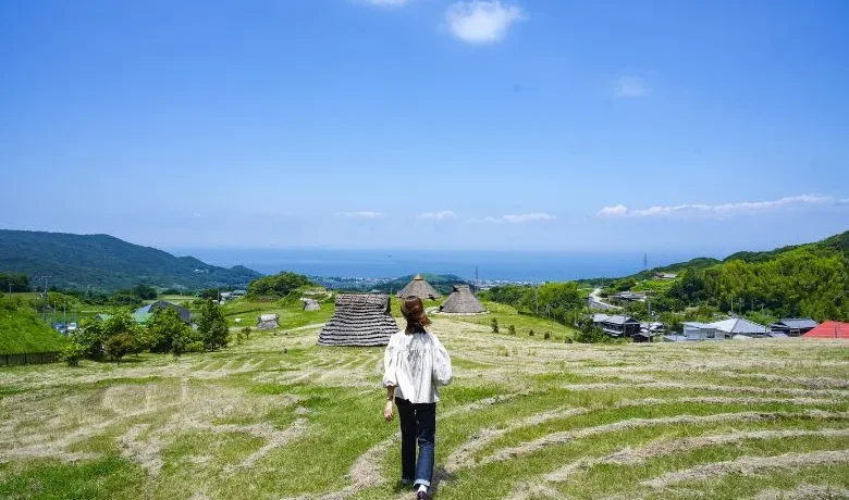 A person walking through a grassy field overlooking the Gossa Kaito Iseki ruins with thatched-roof pit dwellings and the sea in the distance.
