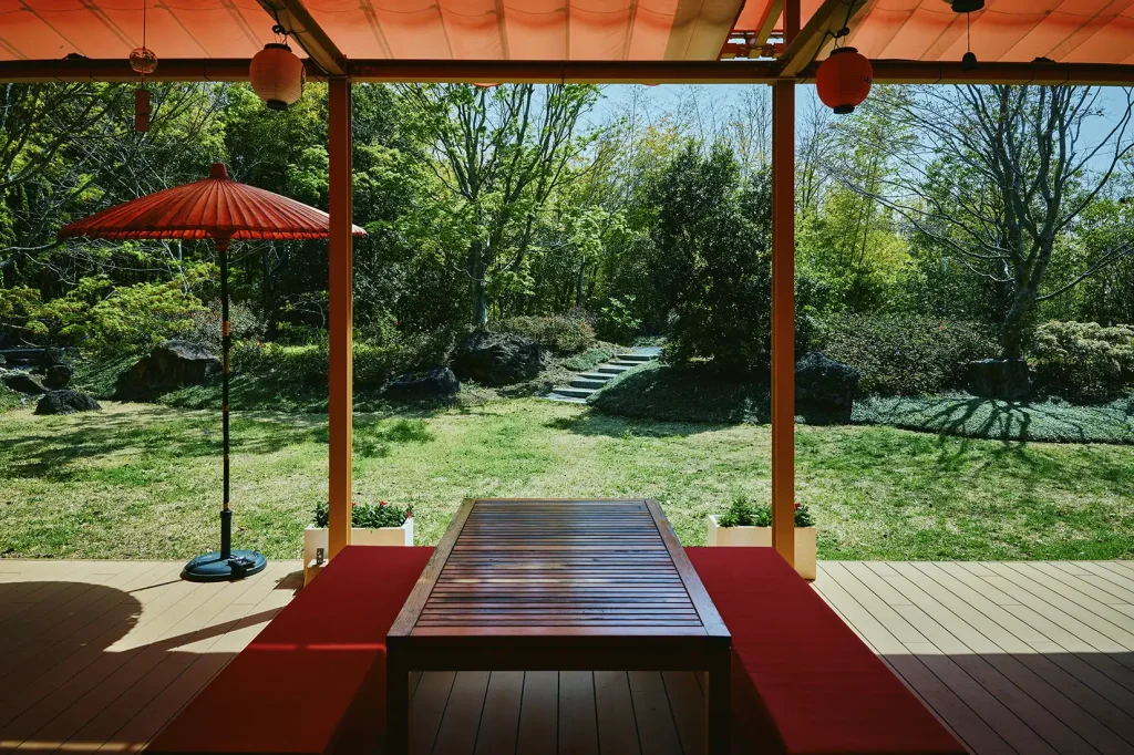 A peaceful outdoor terrace seating area at Aman no Shokutaku, featuring a wooden table and red cushions overlooking a lush Japanese garden.