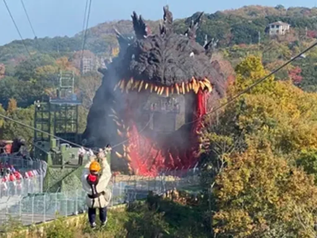 Visitor riding the Godzilla zipline toward the giant Godzilla statue at Nijigen no Mori on Awaji Island, Japan