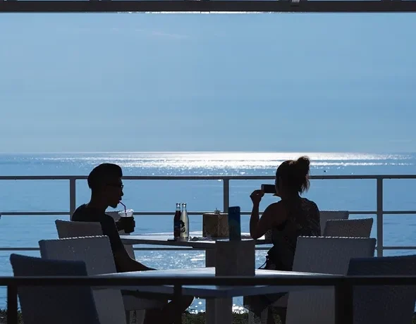 A silhouette of people dining on a terrace at Miele restaurant, overlooking the sparkling blue ocean.